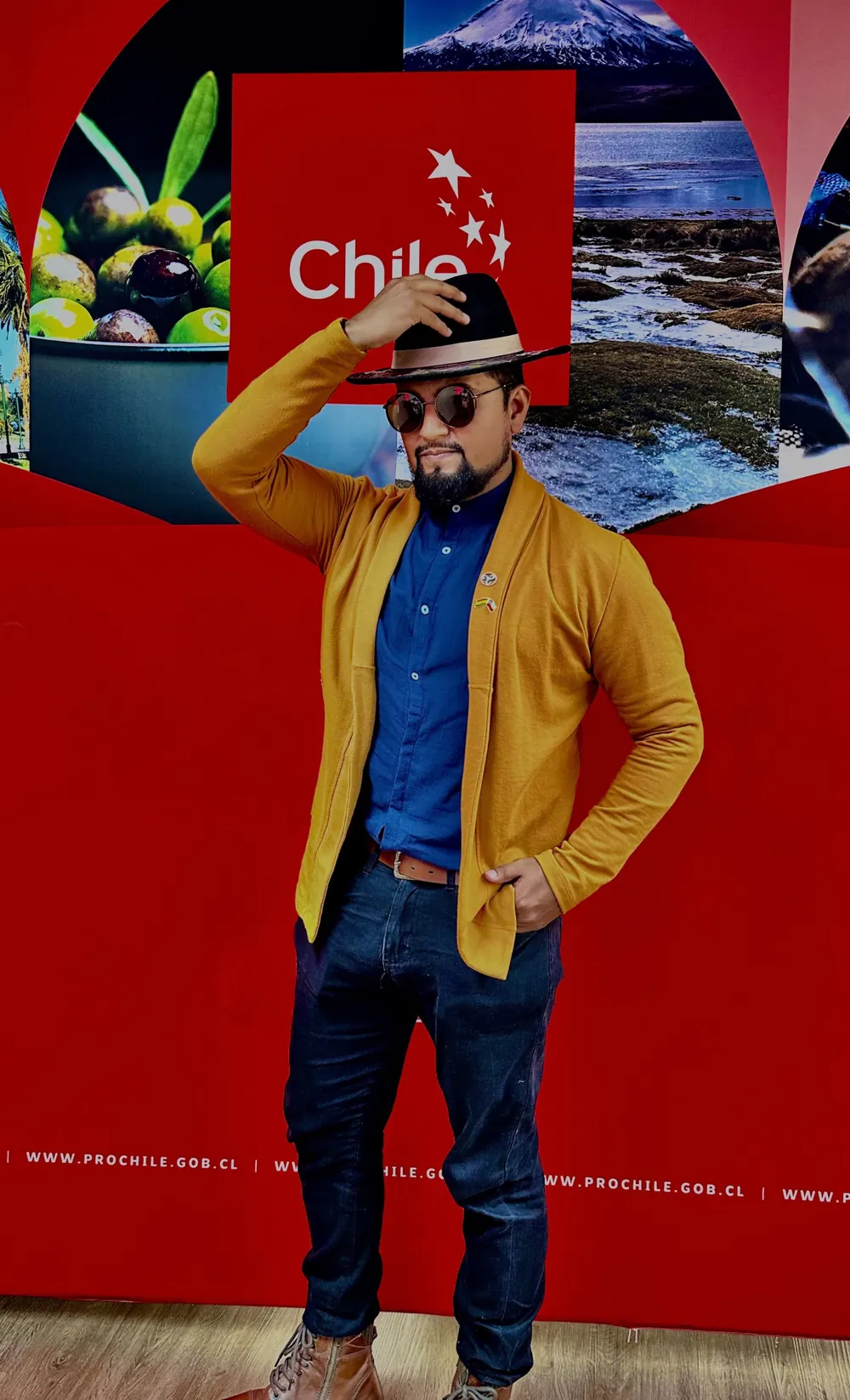 Juan André Hidalgo Ugalde con sombrero, lentes y chaqueta mostaza frente a un panel de ProChile